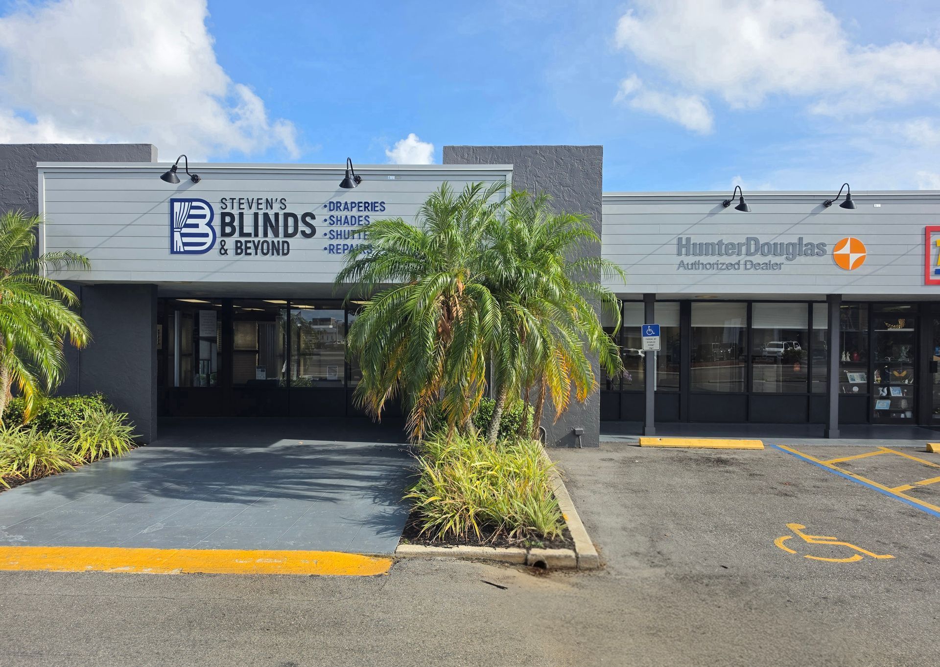 Storefronts in a strip mall, including a Blinds shop and another business, with palm trees and parking spaces.