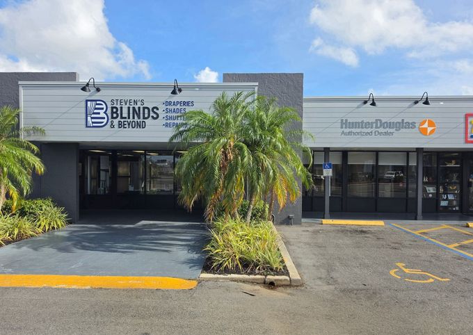 Storefronts in a strip mall, including a Blinds shop and another business, with palm trees and parking spaces.