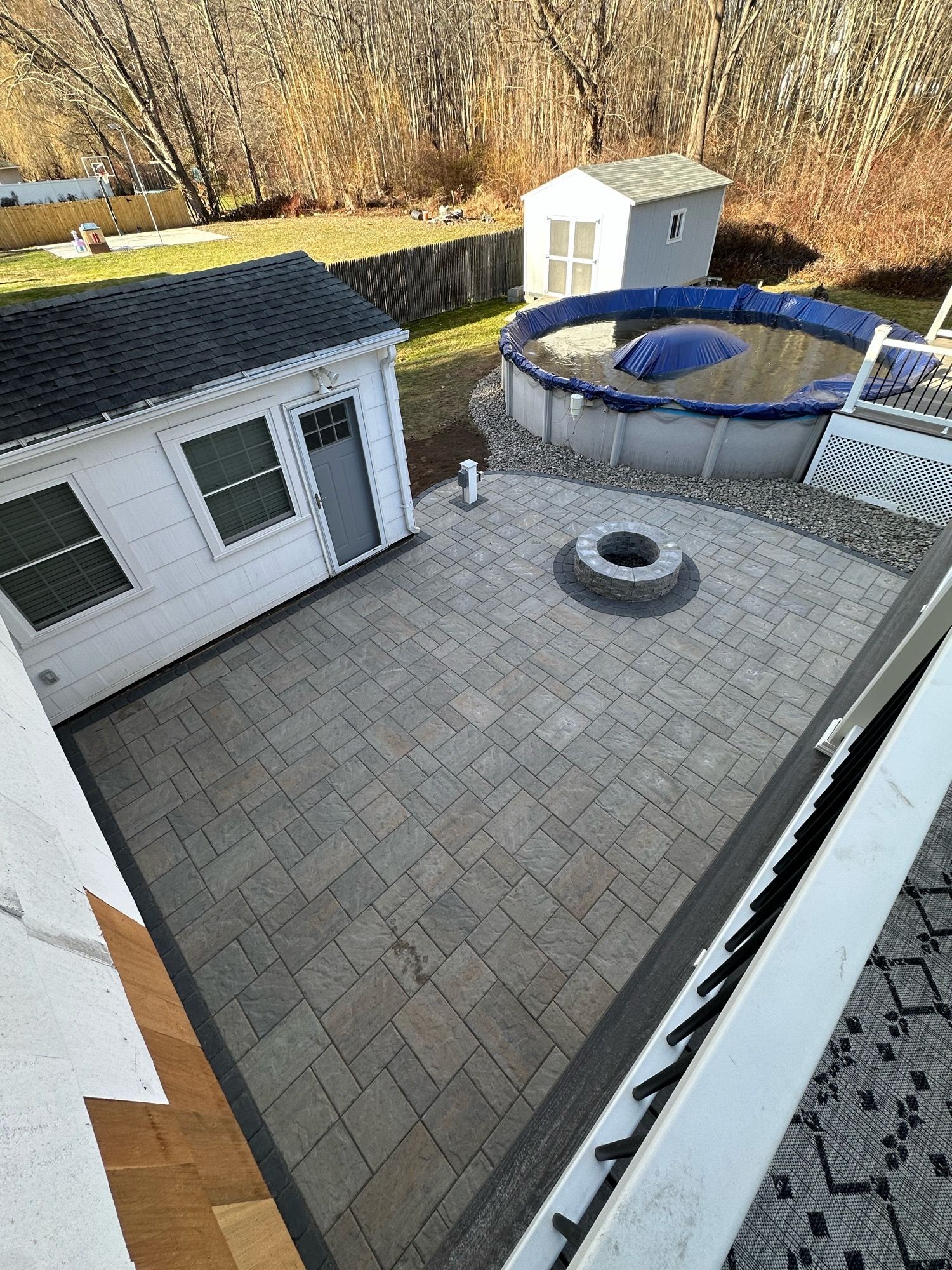 Patio with pavers, fire pit, above-ground pool, and white building in a backyard setting.