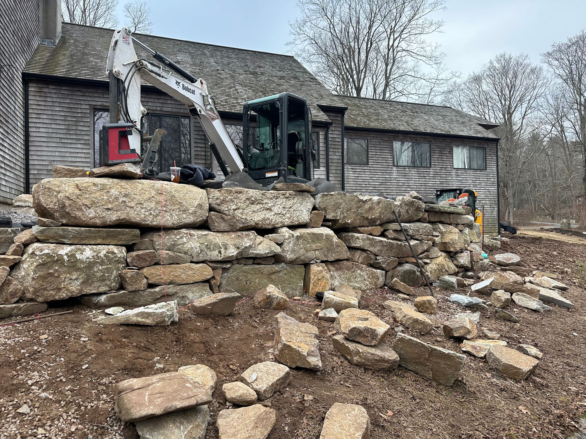 A small excavator building a stone wall near a weathered gray house on a cloudy day.
