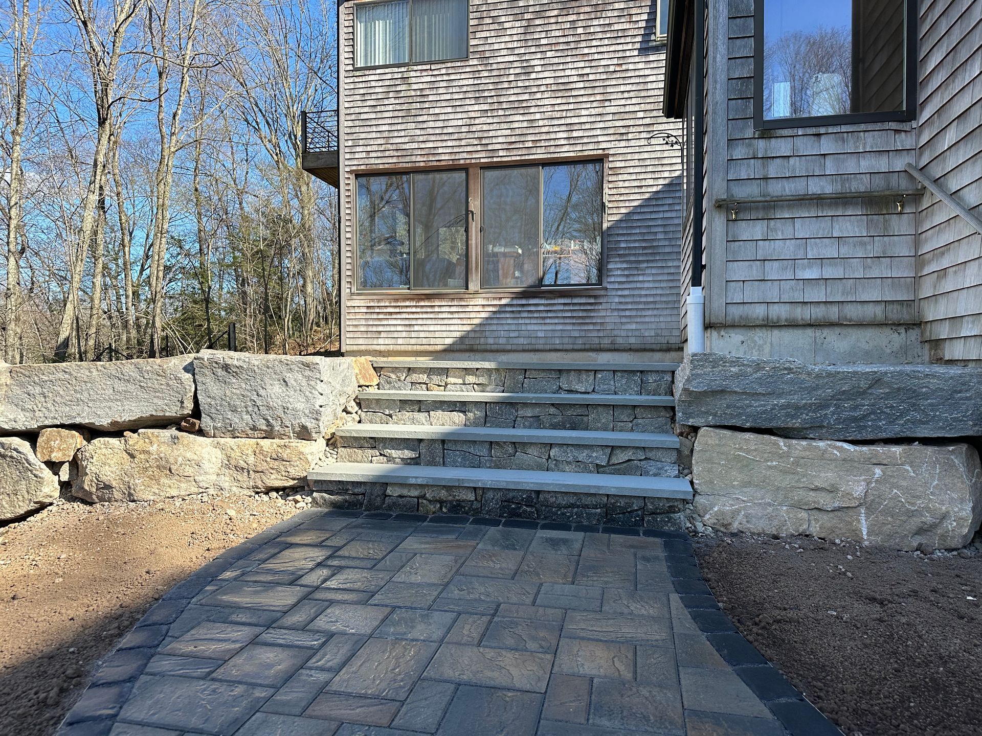 Stone steps leading up to a weathered wooden house with a paved pathway in front, under a sunny sky.