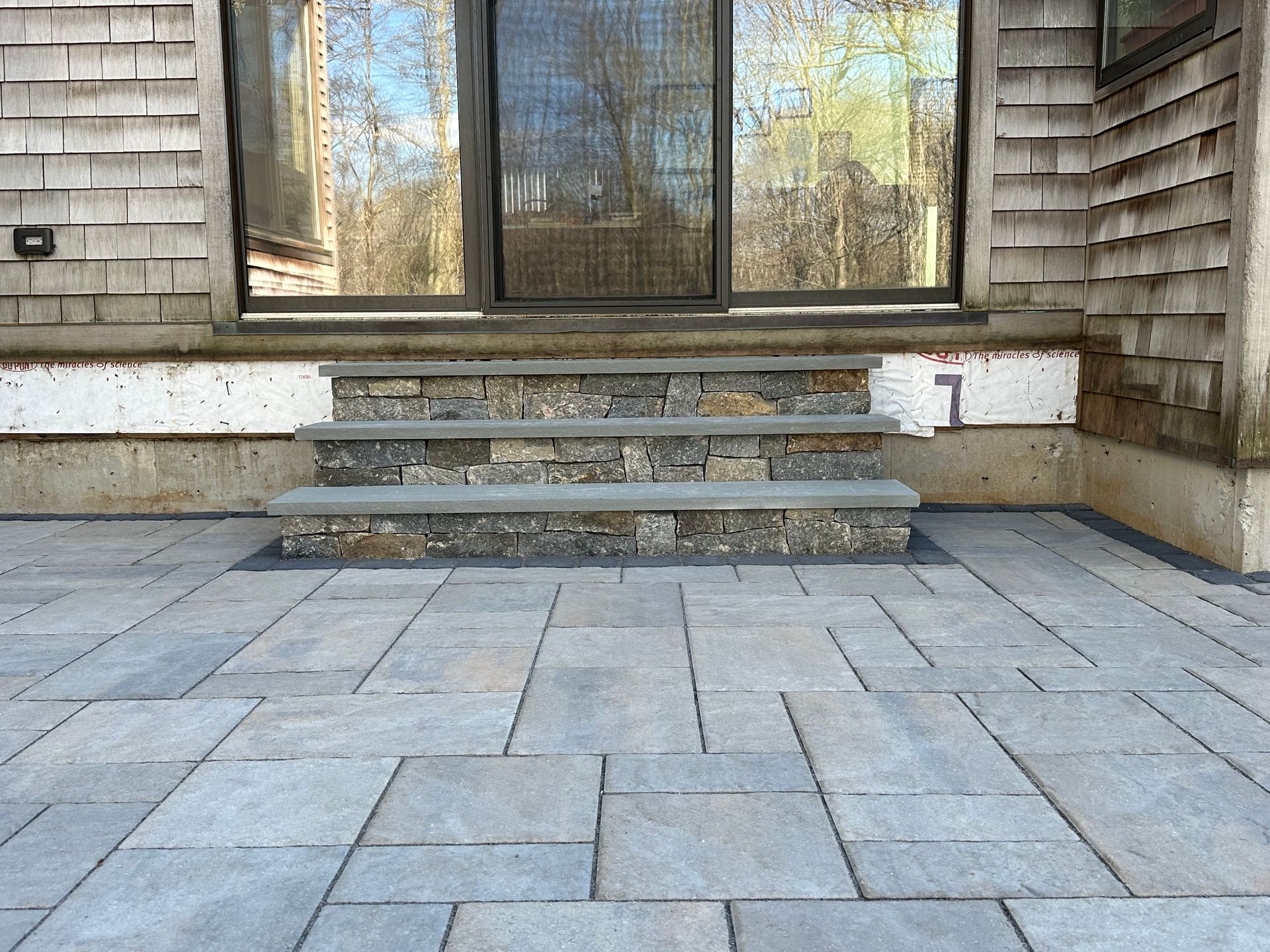 Stone steps leading to a door, set into a paved patio, beneath a wood-sided building.