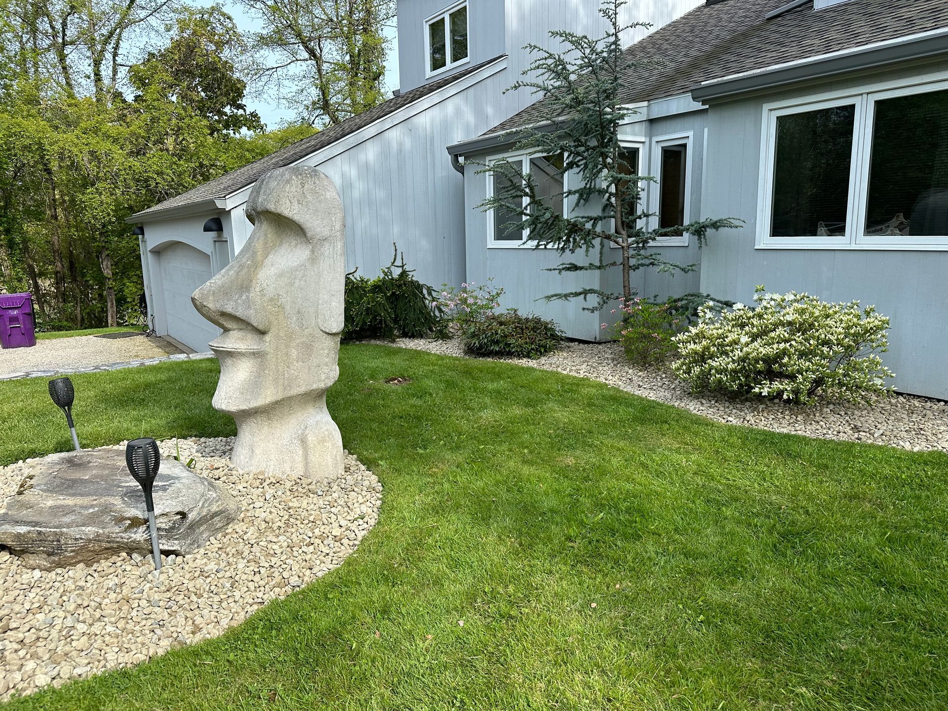 Stone Moai statue in a front yard with green grass, bushes, and a gray house.