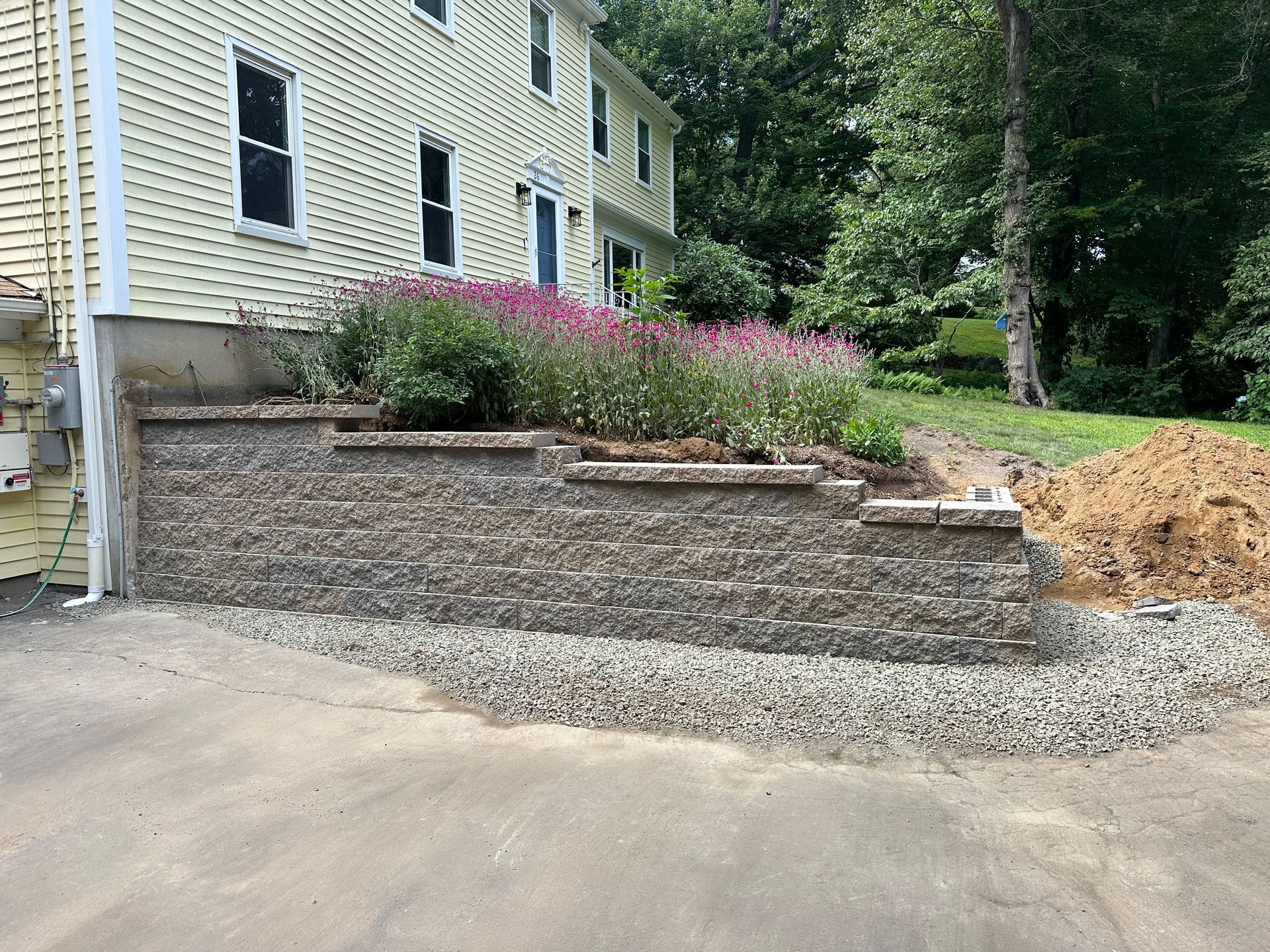 Stone retaining wall with a planted flower bed in front of a yellow house. Gravel at the base.