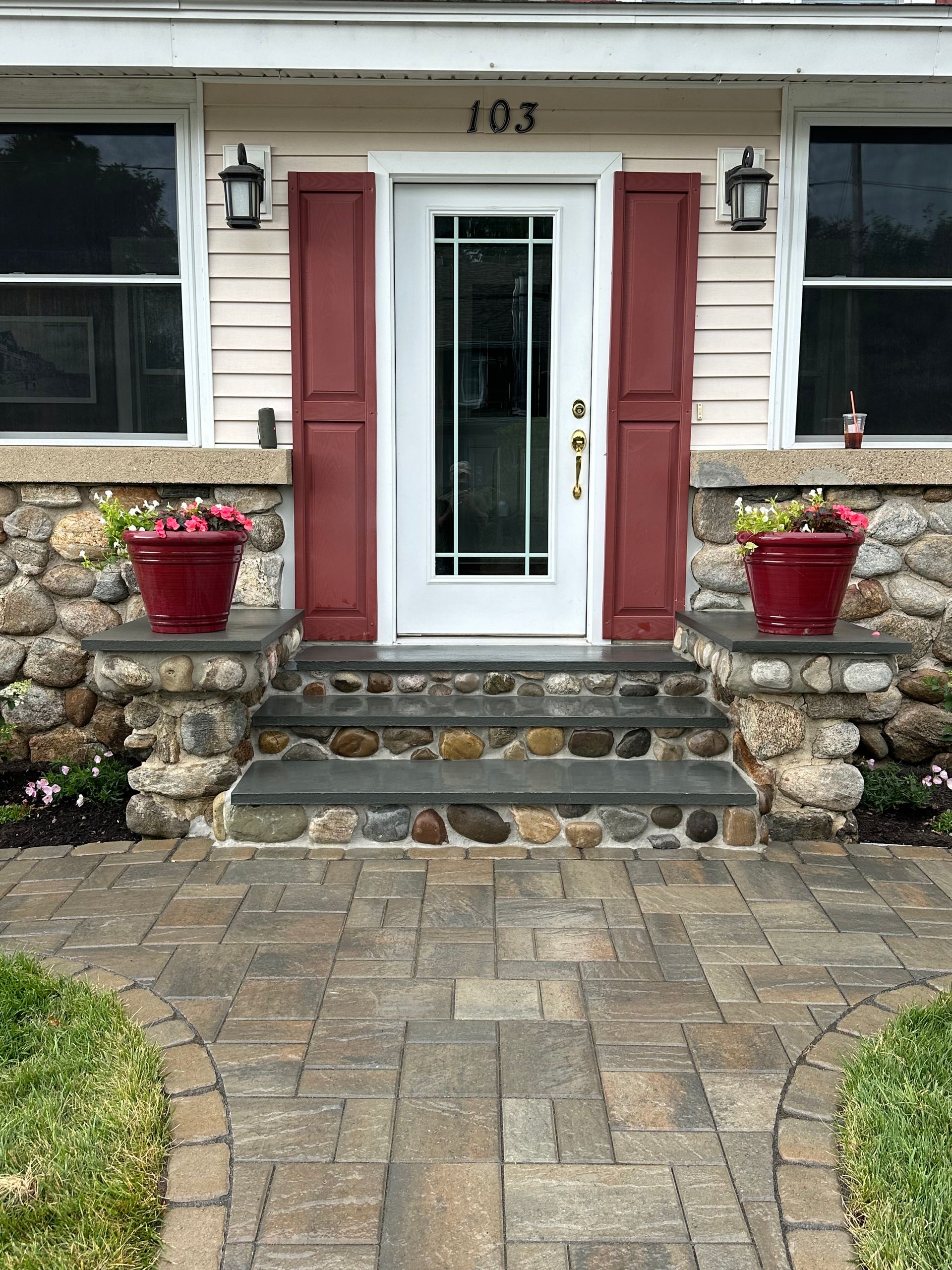 Stone facade home entrance with brick walkway, steps, and red planters and shutters. Door with glass panes.