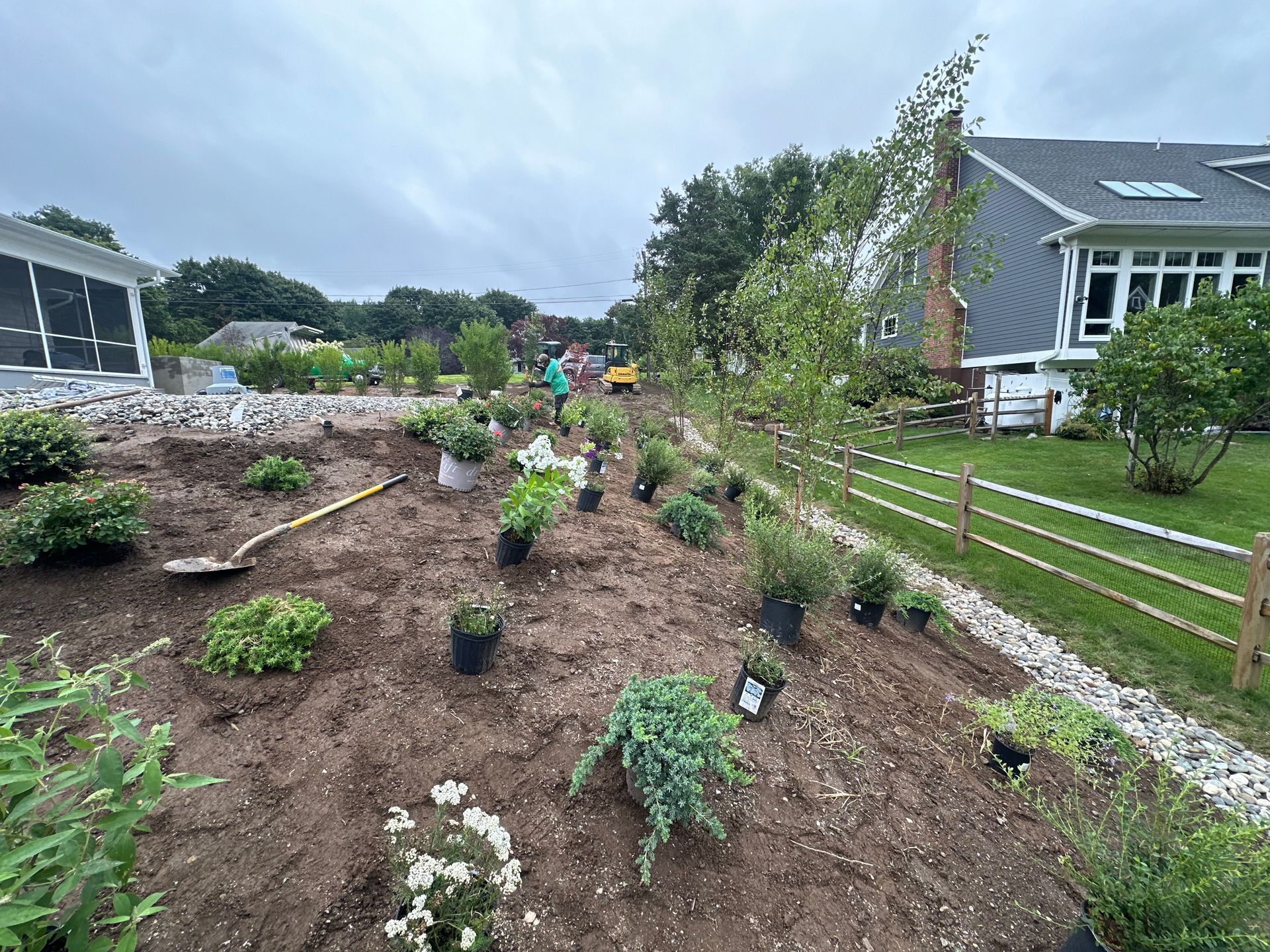 Gardeners planting shrubs in a newly tilled garden bed, near a house with a wooden fence and gravel border.
