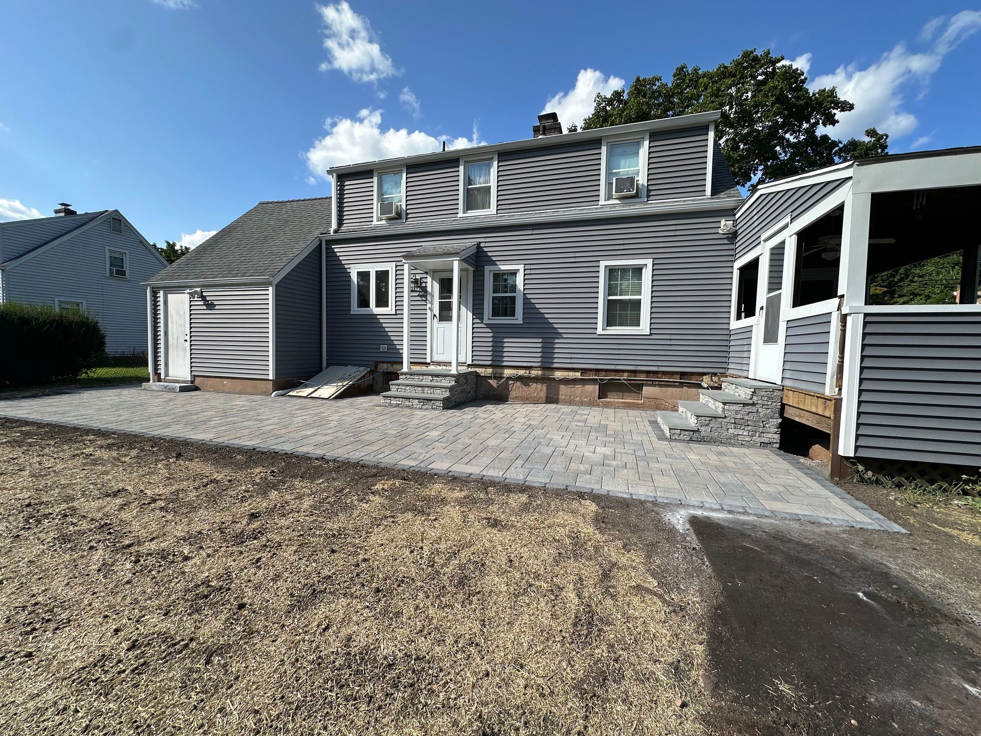 Back of a gray house with a stone patio, under a blue sky. A shed is to the right.