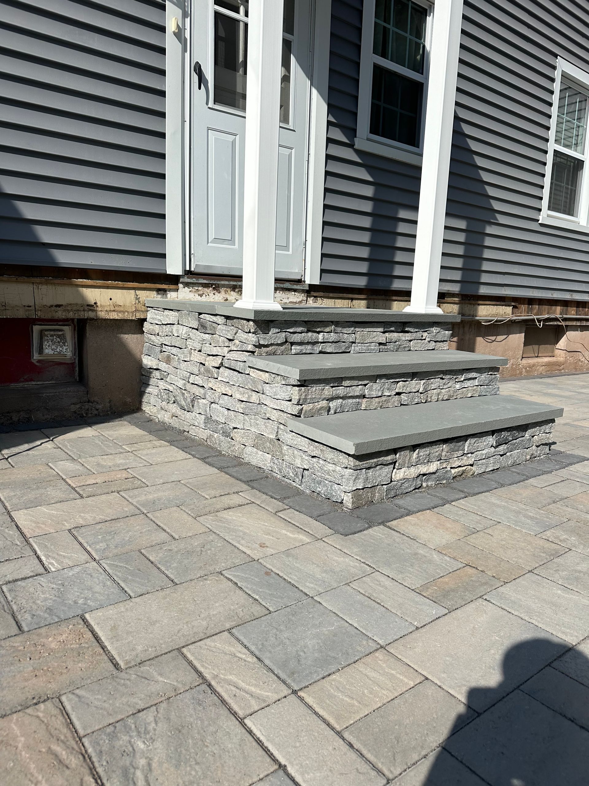 Stone steps leading to a gray door with white pillars, set against a gray house with a stone patio.