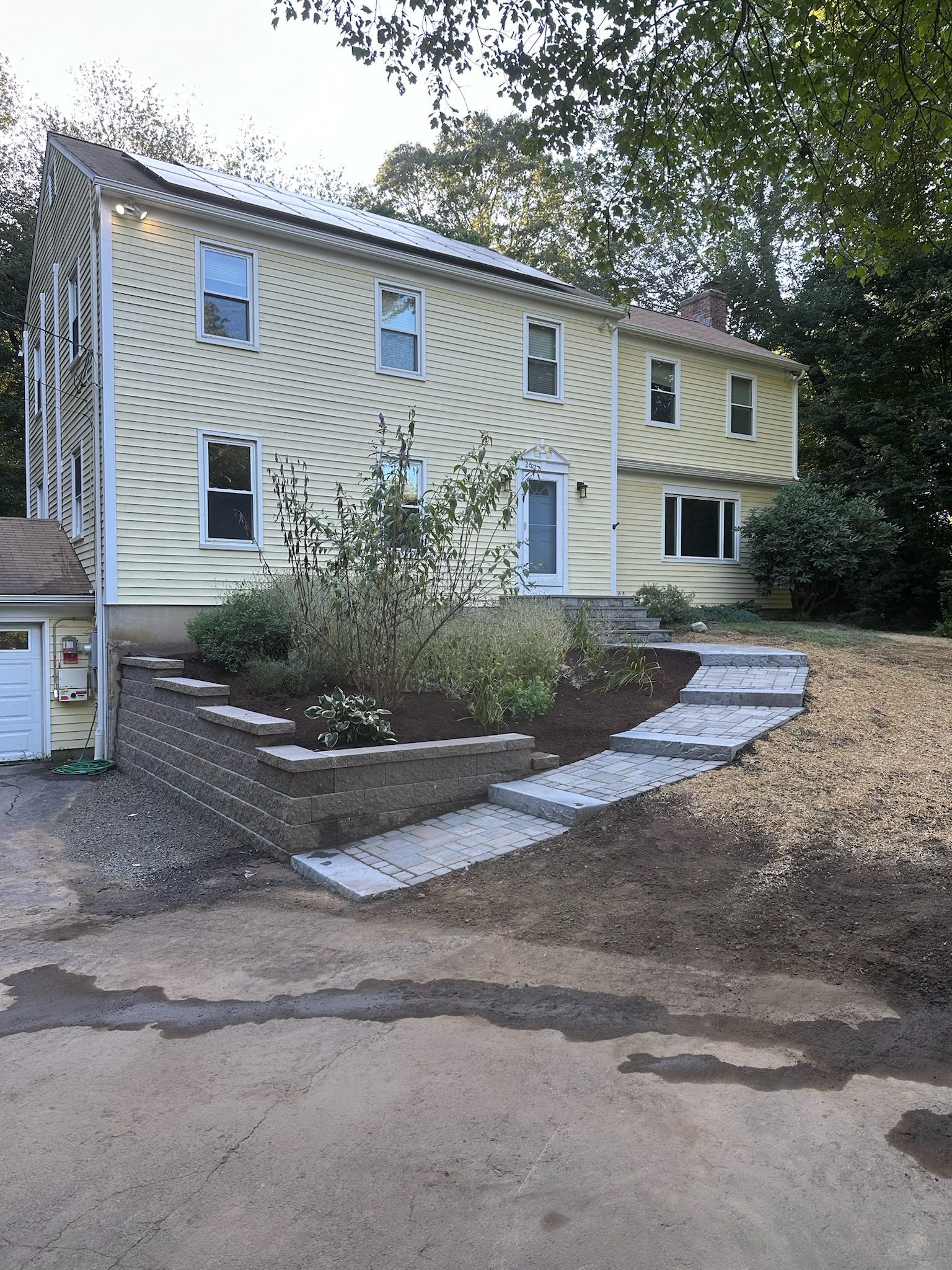 Two-story yellow house with paved steps leading to the front door, landscaped garden bed with mulch.