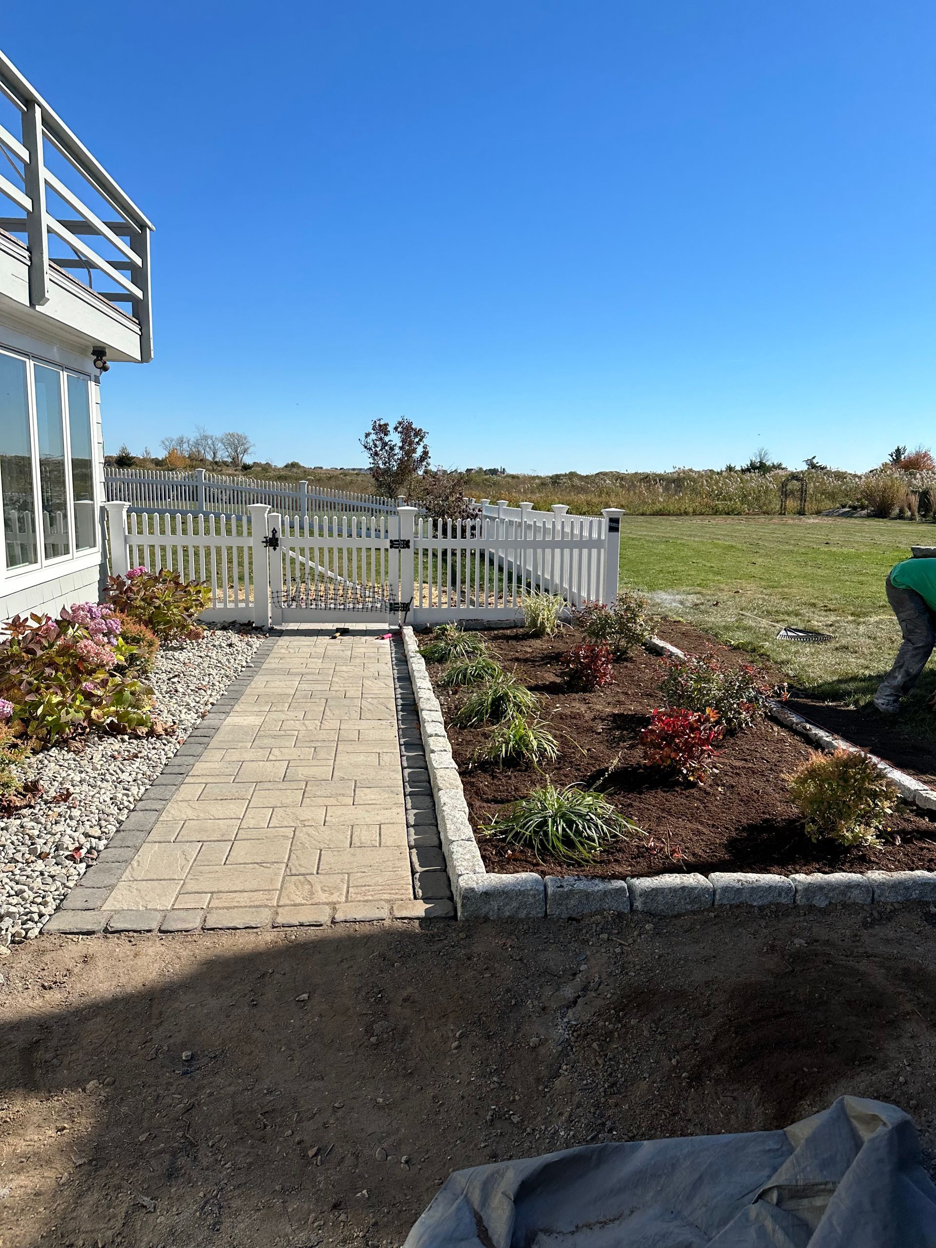 Stone path leads to a white picket fence, garden with flowers, next to a building on a sunny day.
