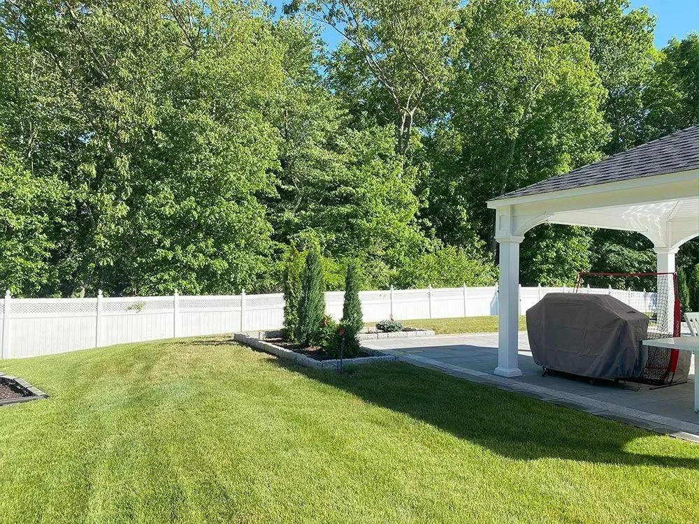 Lush green backyard with a white fence, patio, and gazebo. Trees in the background, a grill covered.