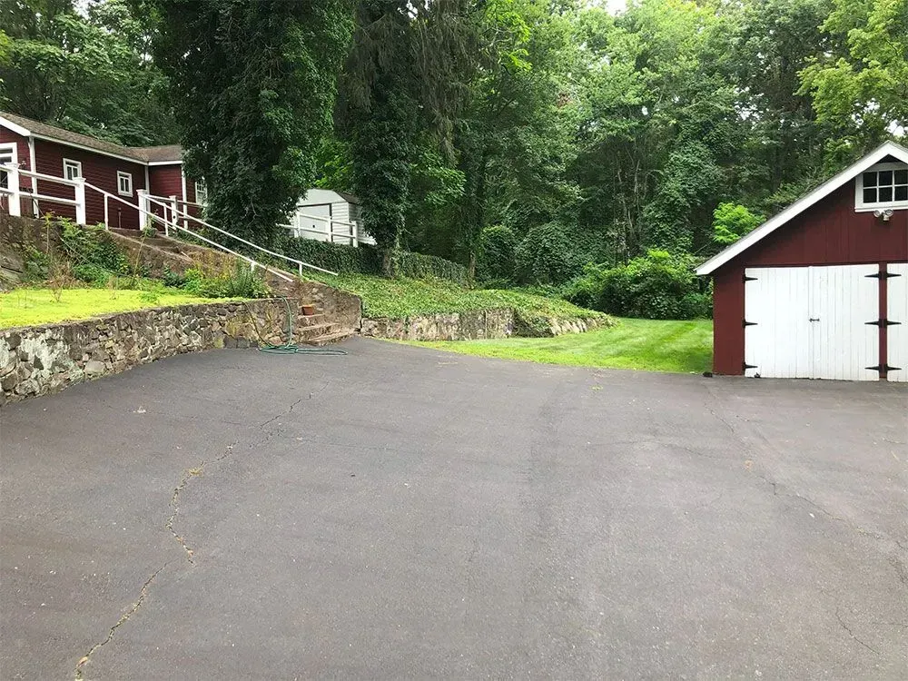 Asphalt driveway in front of a red garage and house, bordered by a stone wall and lush greenery.