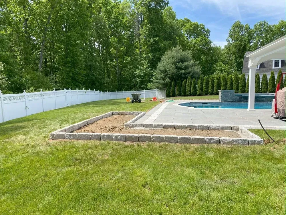 Backyard with a raised garden bed, patio, pool, and white fence. Green grass and trees in the background.