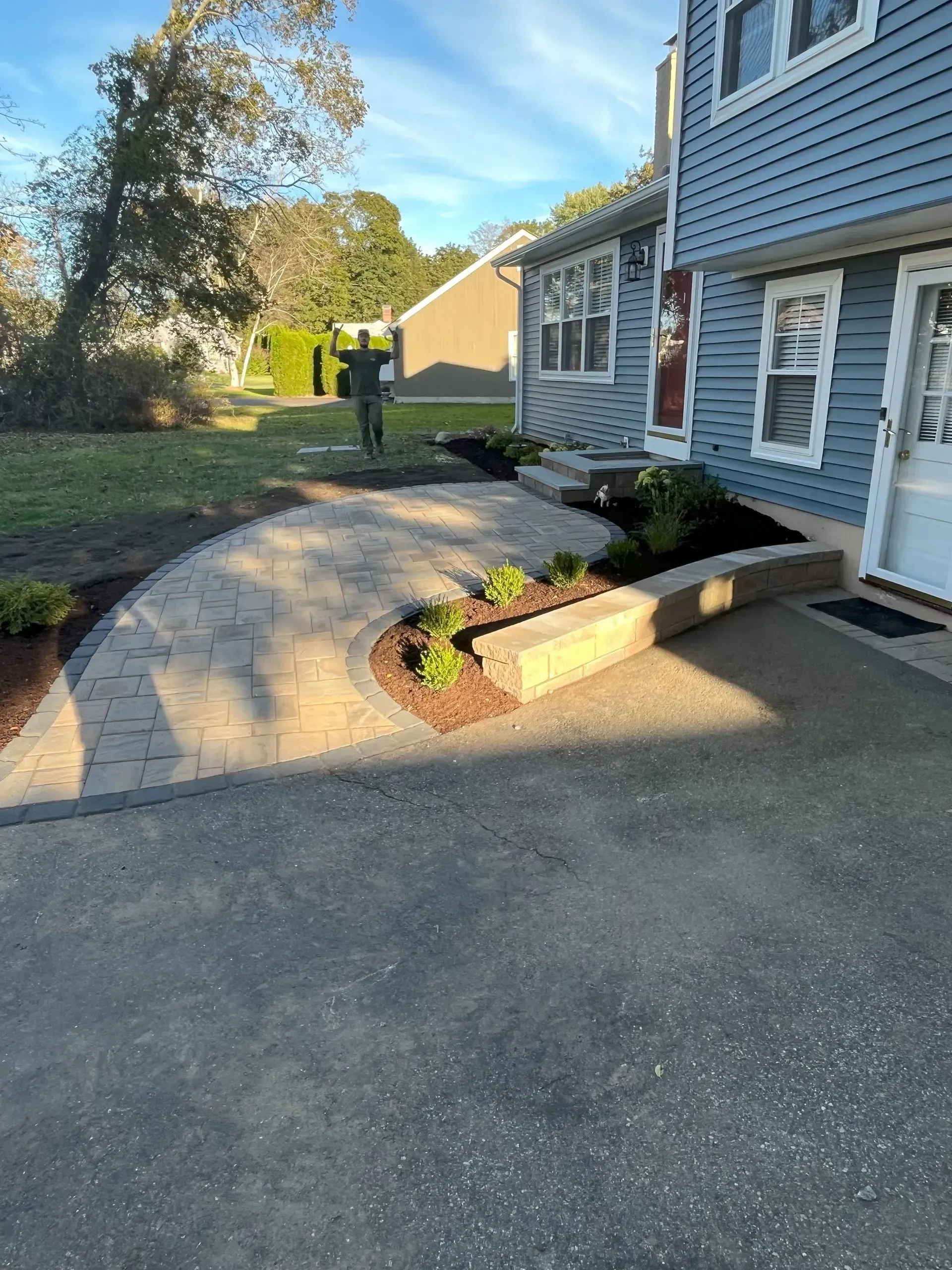 Paver walkway with a raised flower bed in front of a blue-sided house with a door.