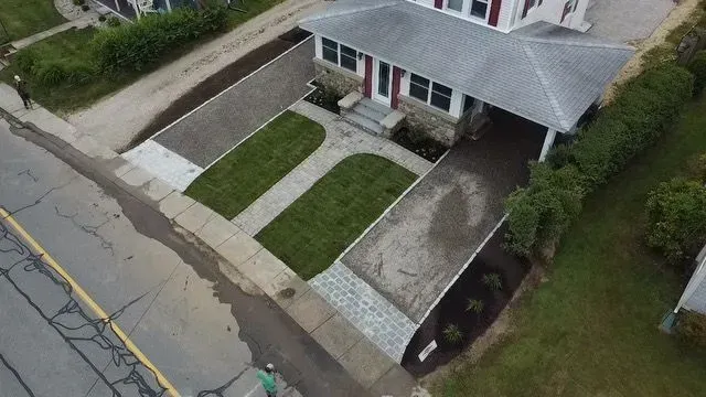 Aerial view of a house with a gray roof and driveway.  Green grass and concrete walkways are in the front yard.