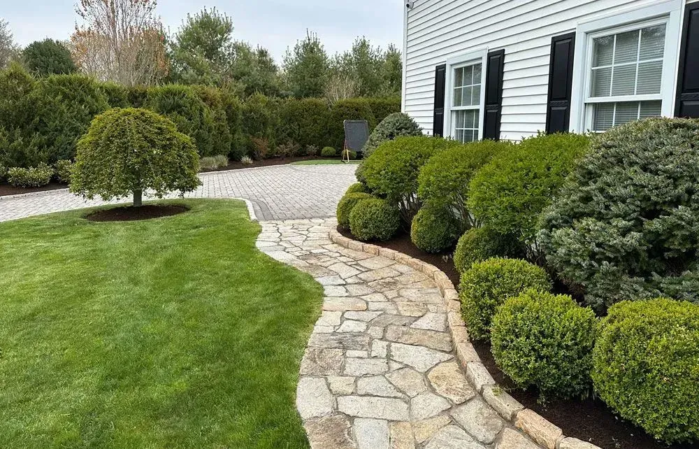 Stone path curves through manicured lawn and garden, leading to a white house with black shutters.
