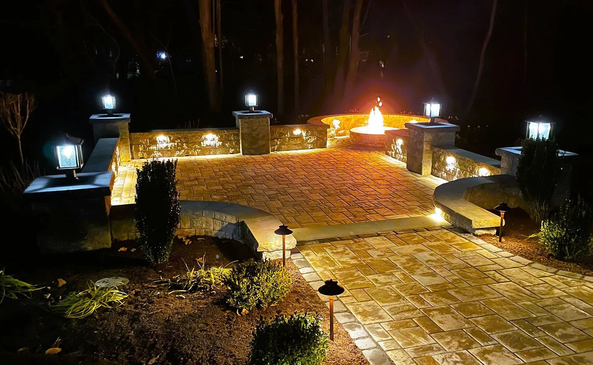 A lit stone patio at night with a fire pit, illuminated by lamps and landscape lights.