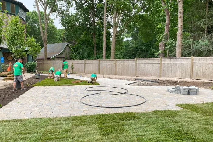 Landscapers installing a gravel patio in a yard with fresh sod. Several workers are present near a building and trees.