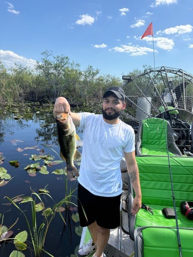 A man holding a fish on an airboat in a swamp, sunny day.