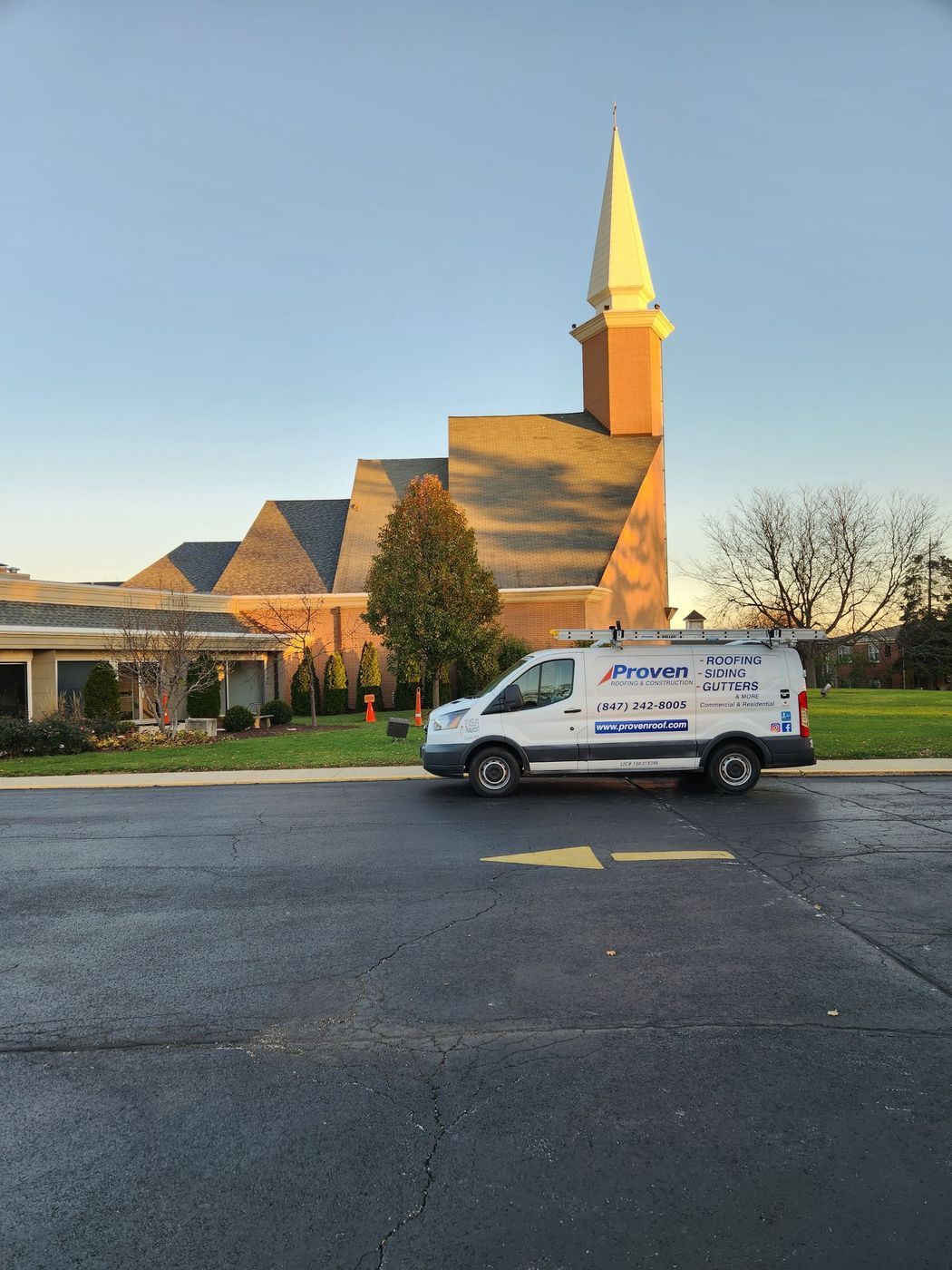 A white van parked on pavement in front of a church with a tall steeple on a sunny day.