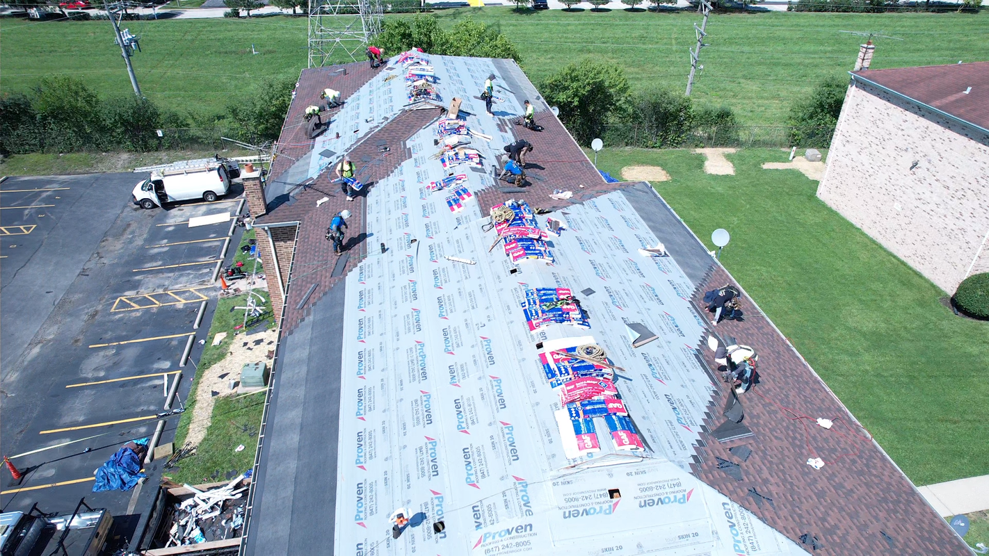 Roofers working on a building, applying shingles. Dark and light brown roof, sunny day, with green lawn.