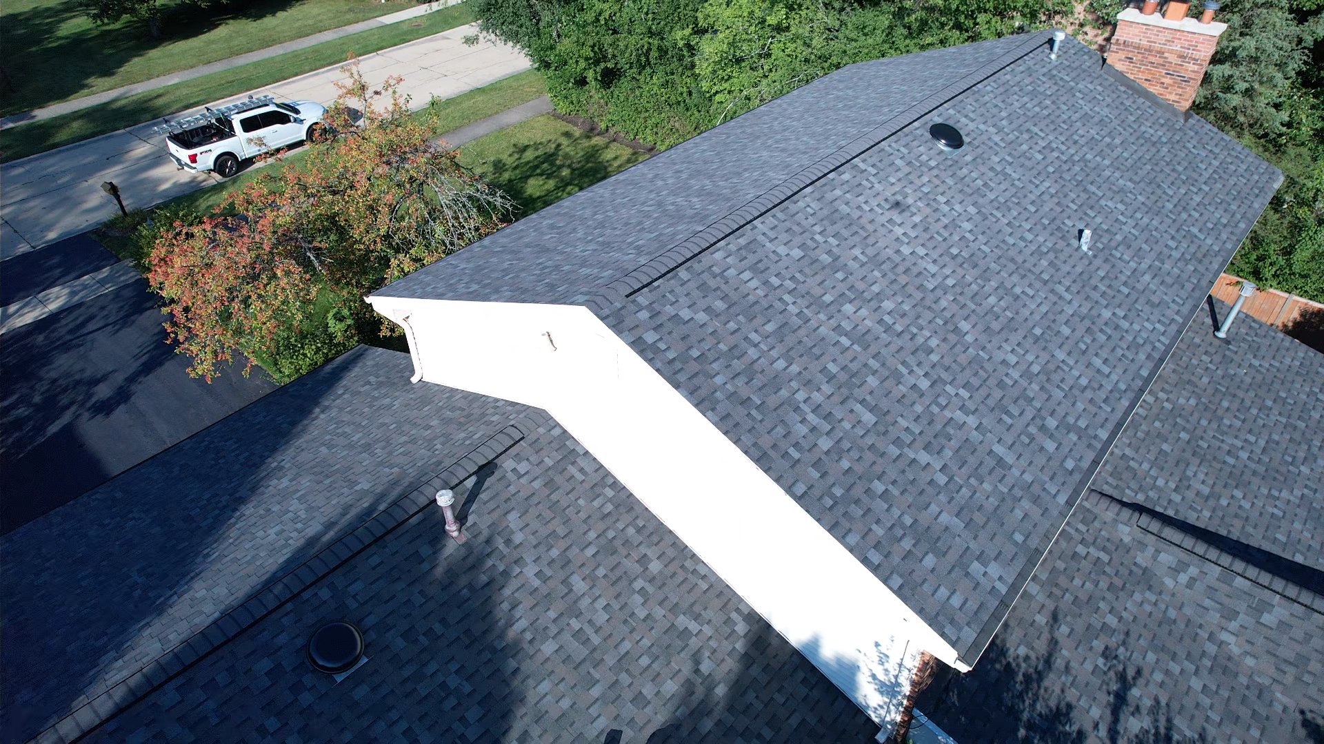 Overhead view of a house roof covered in dark shingles. White wall and a chimney are visible. A truck and trees are in the background.