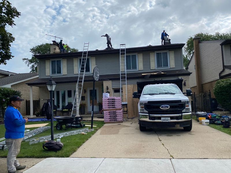Roofers on a house with a truck in the driveway, supplies, and a person observing.
