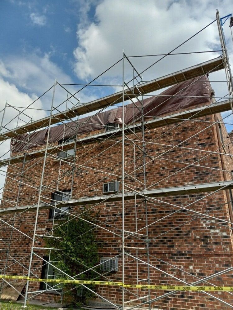 Scaffolding covers brick building, with tarp on top level. Yellow caution tape in foreground, cloudy sky above.