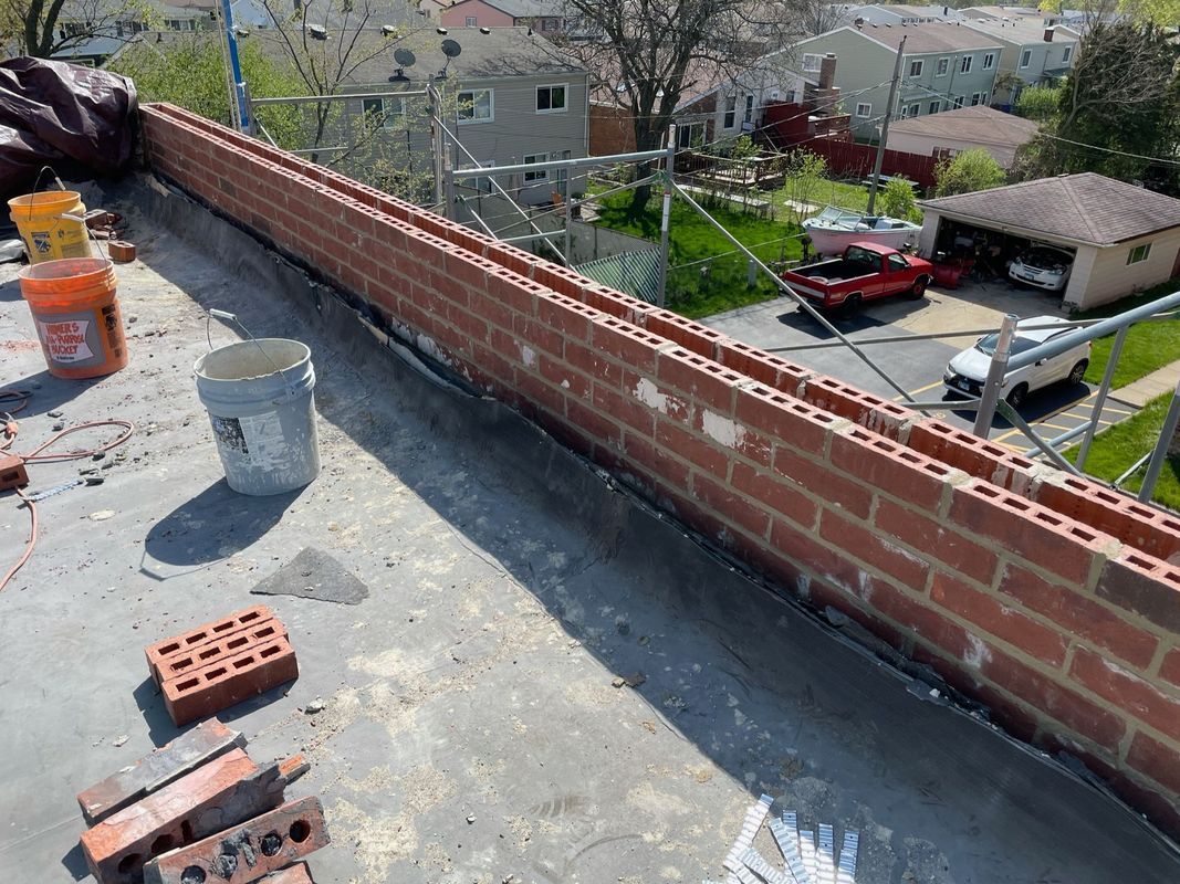 Brick wall construction on a flat roof, with bricks, mortar, and tools visible. Bright daylight.