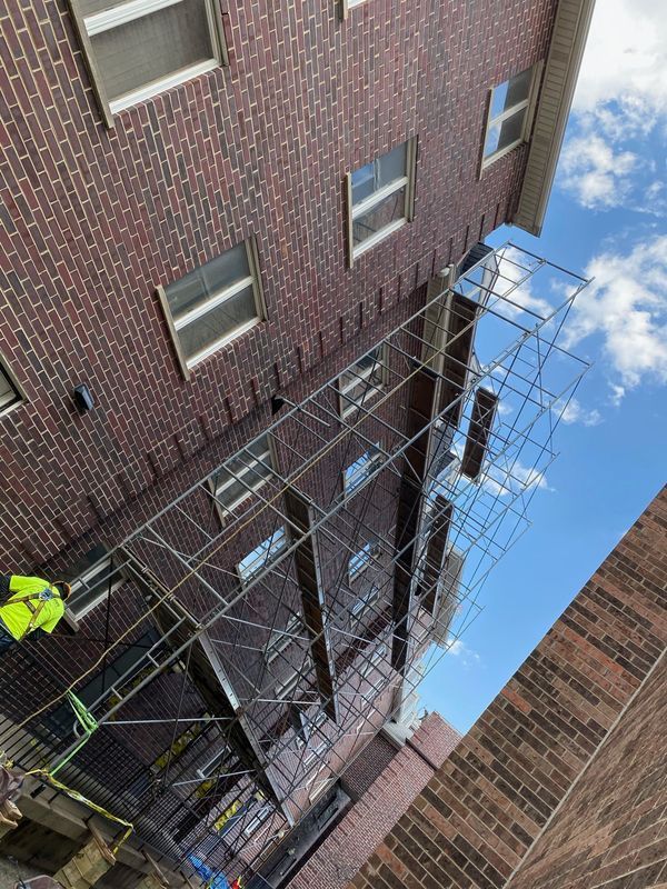 Scaffolding alongside a brick building; worker in yellow vest. Blue sky in the background.
