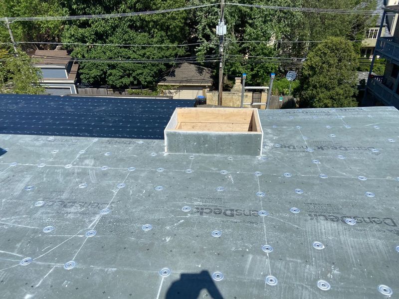 Rooftop with a square wooden structure and surrounding grey underlayment, trees in background.