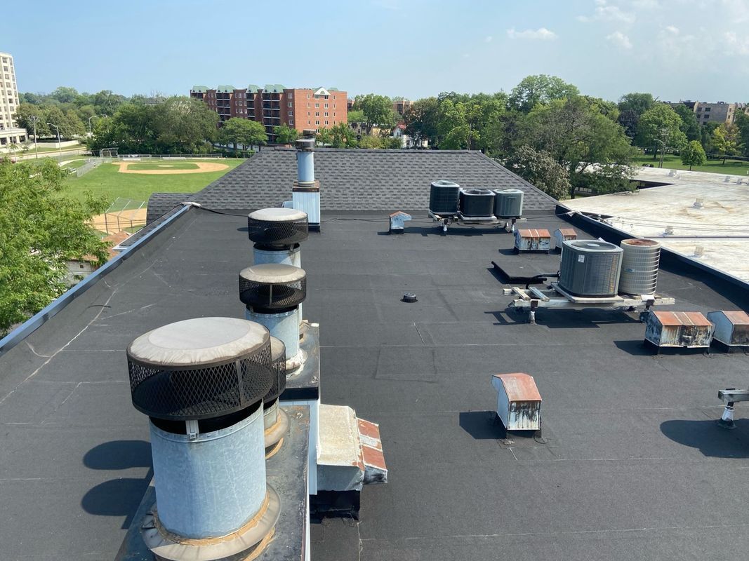 Flat, black rooftop with vents, machinery, and a view of buildings and trees.