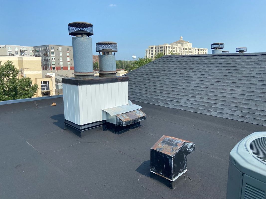 Rooftop with chimneys and air conditioning units under a blue sky, buildings in the background.