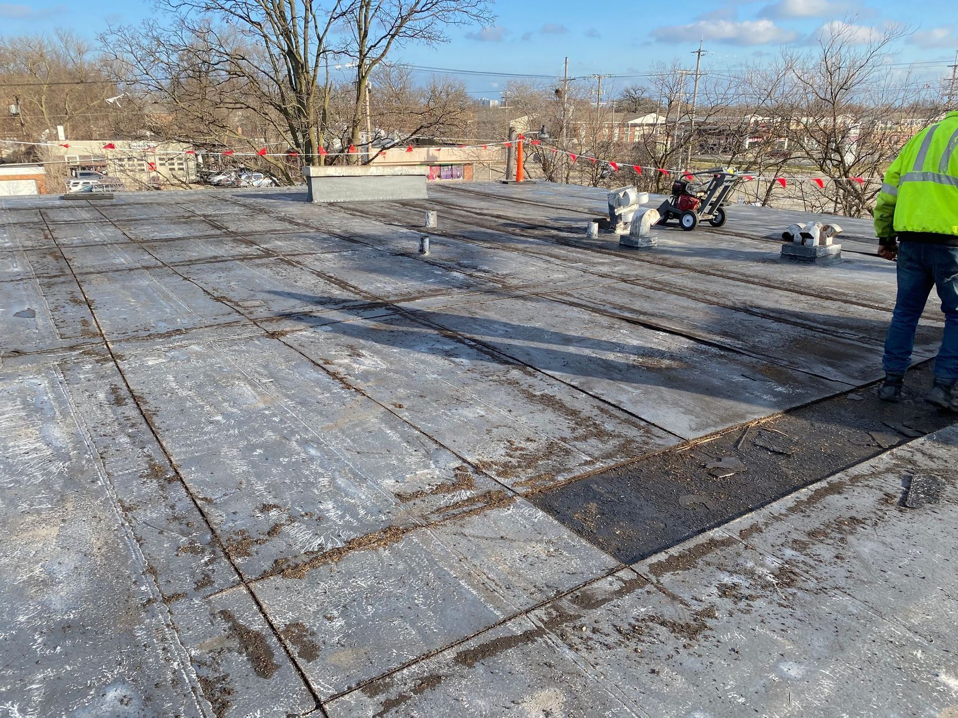 Workers on a flat roof cutting and preparing for repairs; a person in a safety vest stands to the right.