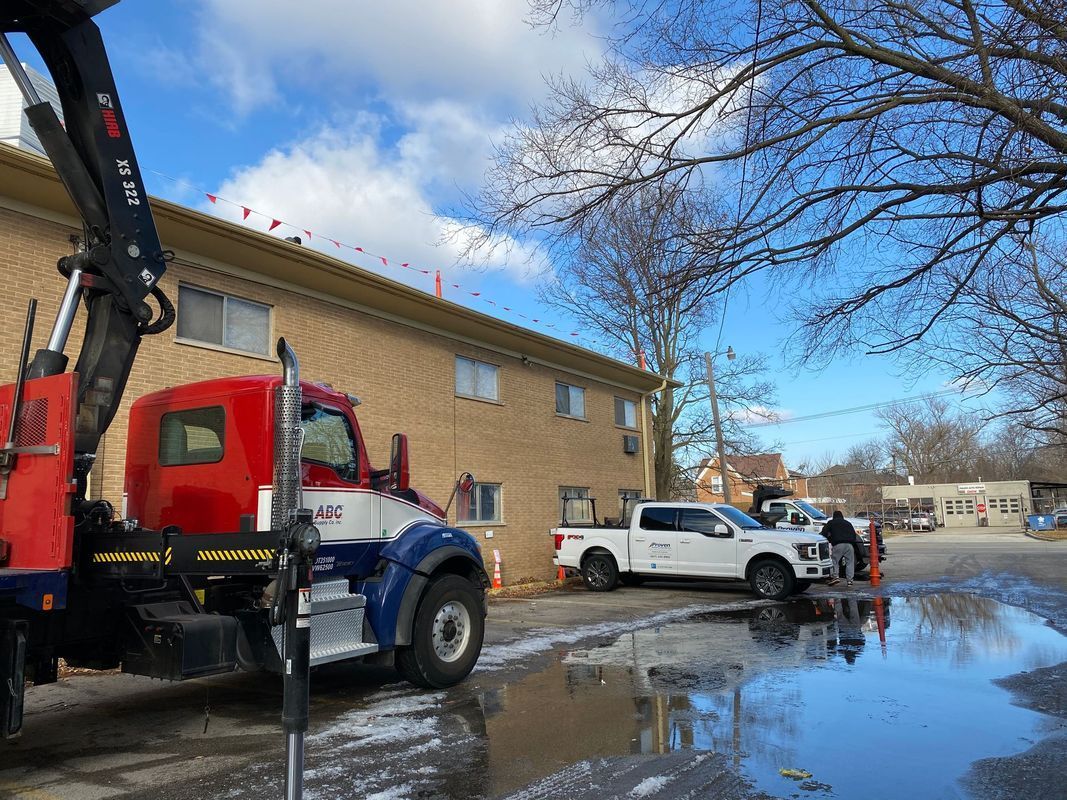 A red, white, and blue truck with an articulated arm parked near a building. A puddle reflects the sky.
