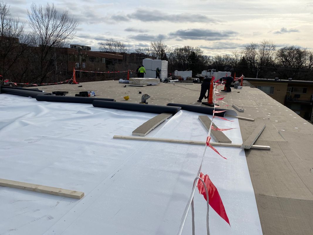 Workers installing a white roof membrane on a flat rooftop under an overcast sky.