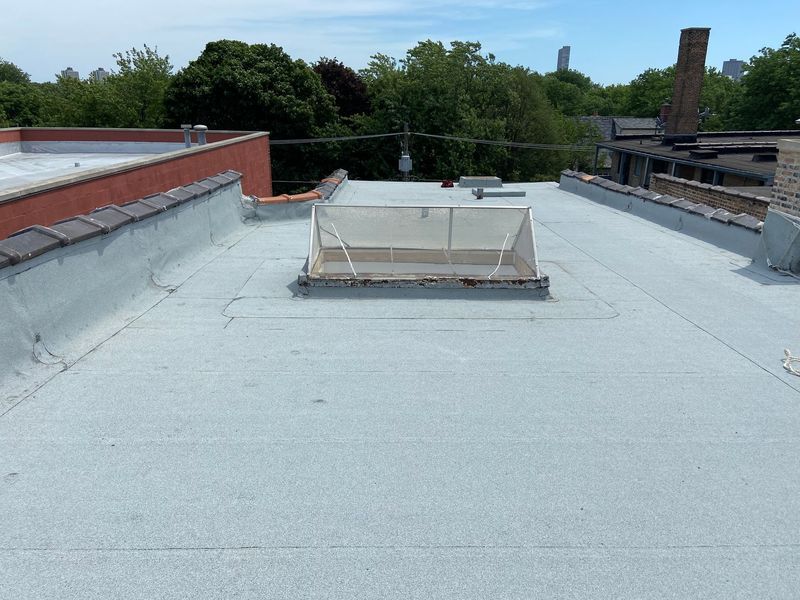 Flat, light-gray rooftop with a skylight, surrounded by trees and buildings on a sunny day.