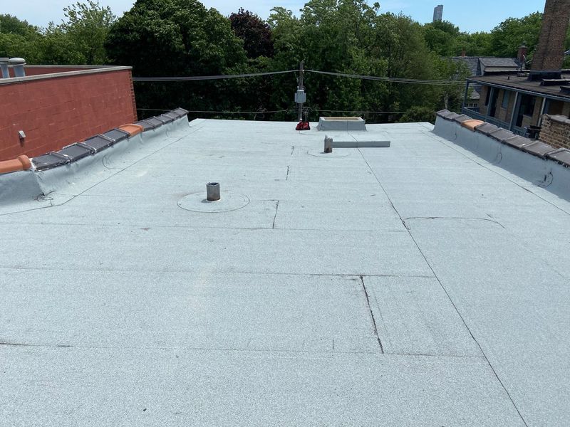 Flat, light-colored roof with various vents and seams, surrounded by a low wall. Trees and sky in the background.