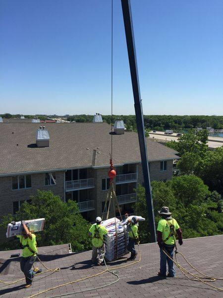 Construction workers on a rooftop use a crane to lift materials. Bright blue sky, sunny day.