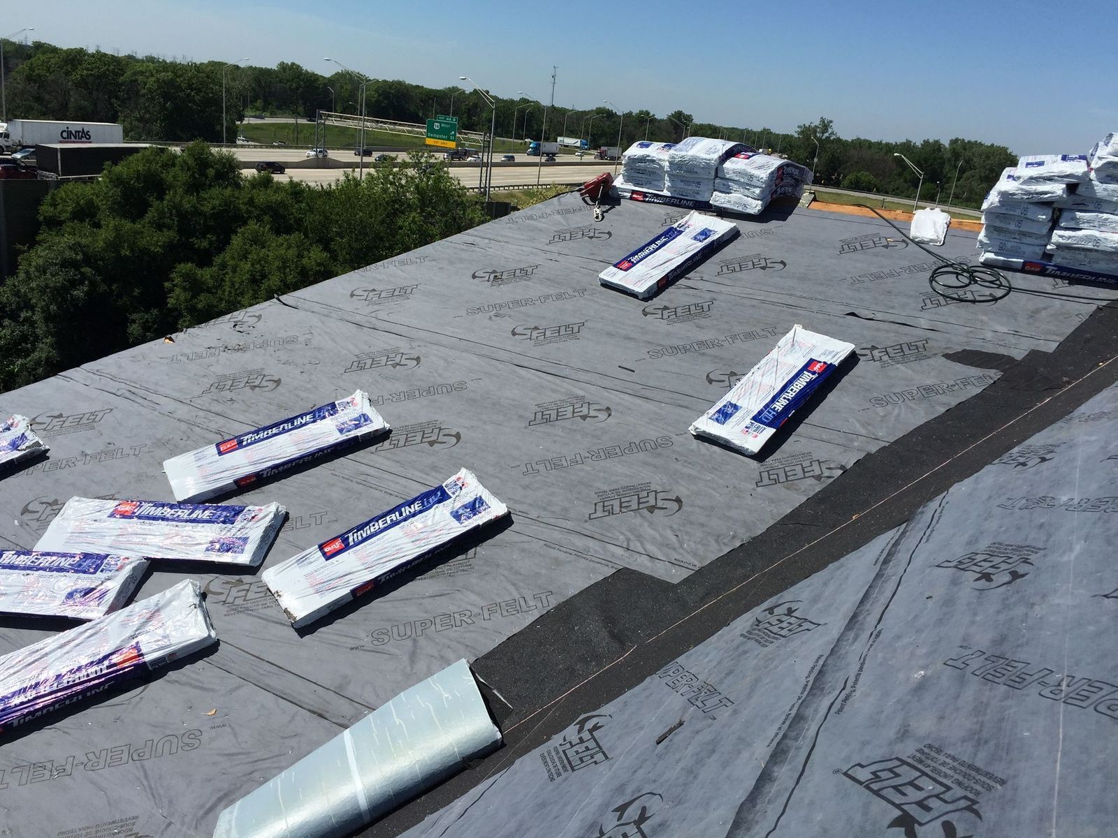 Rooftop under construction with roofing materials scattered. Black underlayment with packages, highway in background.