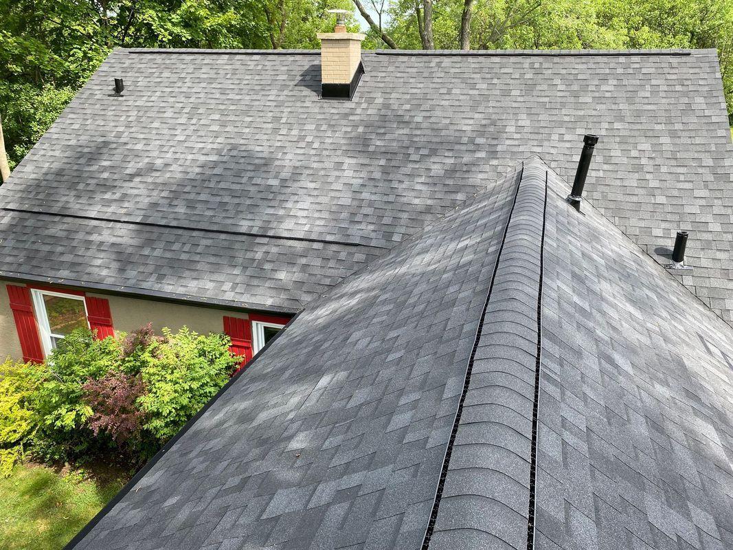 Gray asphalt shingle roof on a house, angled view. Chimney, vents visible. Green trees and red shutters.