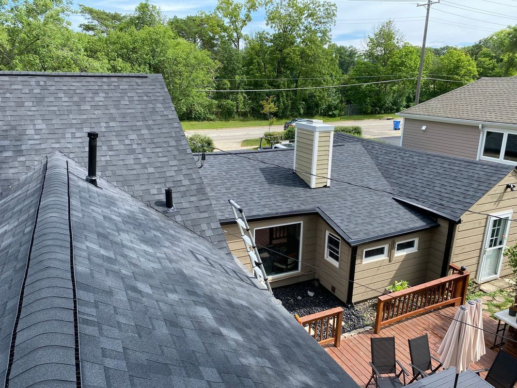 Dark gray shingle roofs of multiple houses with a light brown exterior and trees in the background.