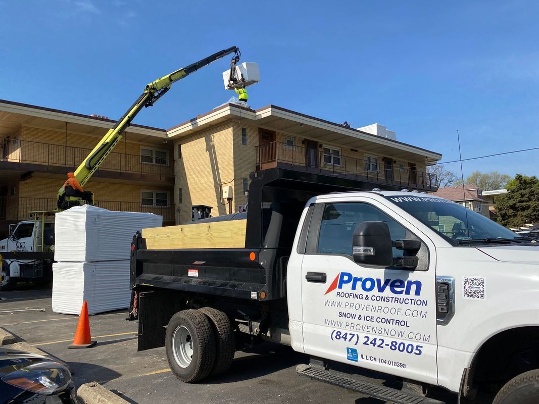Truck with crane lifts materials to a rooftop; worker on roof, another truck nearby.
