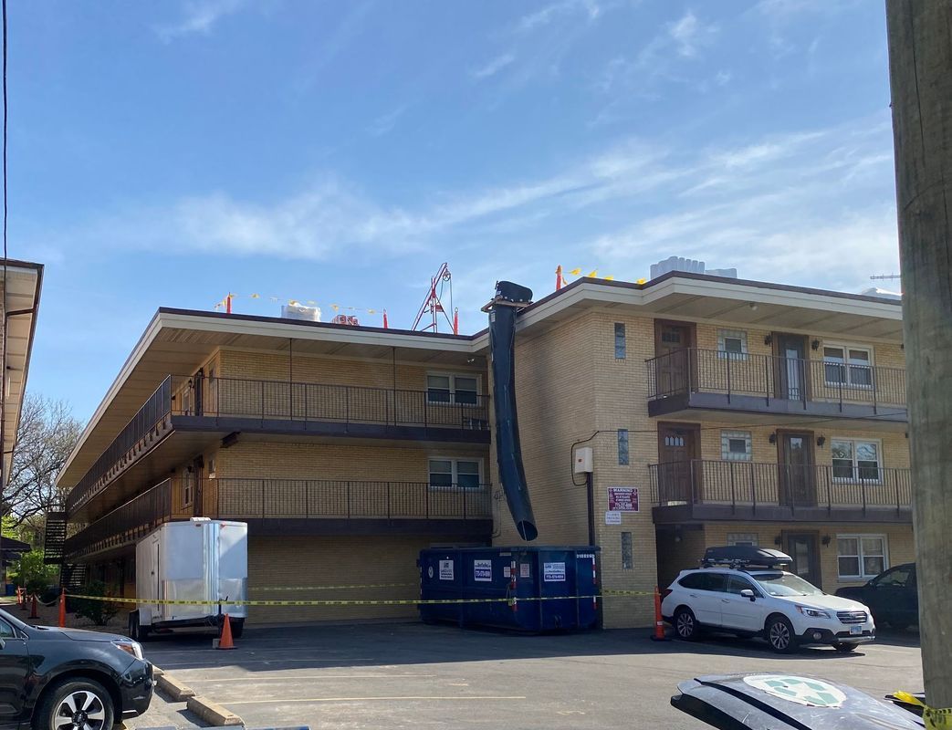 Two-story brick motel with a black chute extending from the roof to a dumpster, with vehicles parked in front.