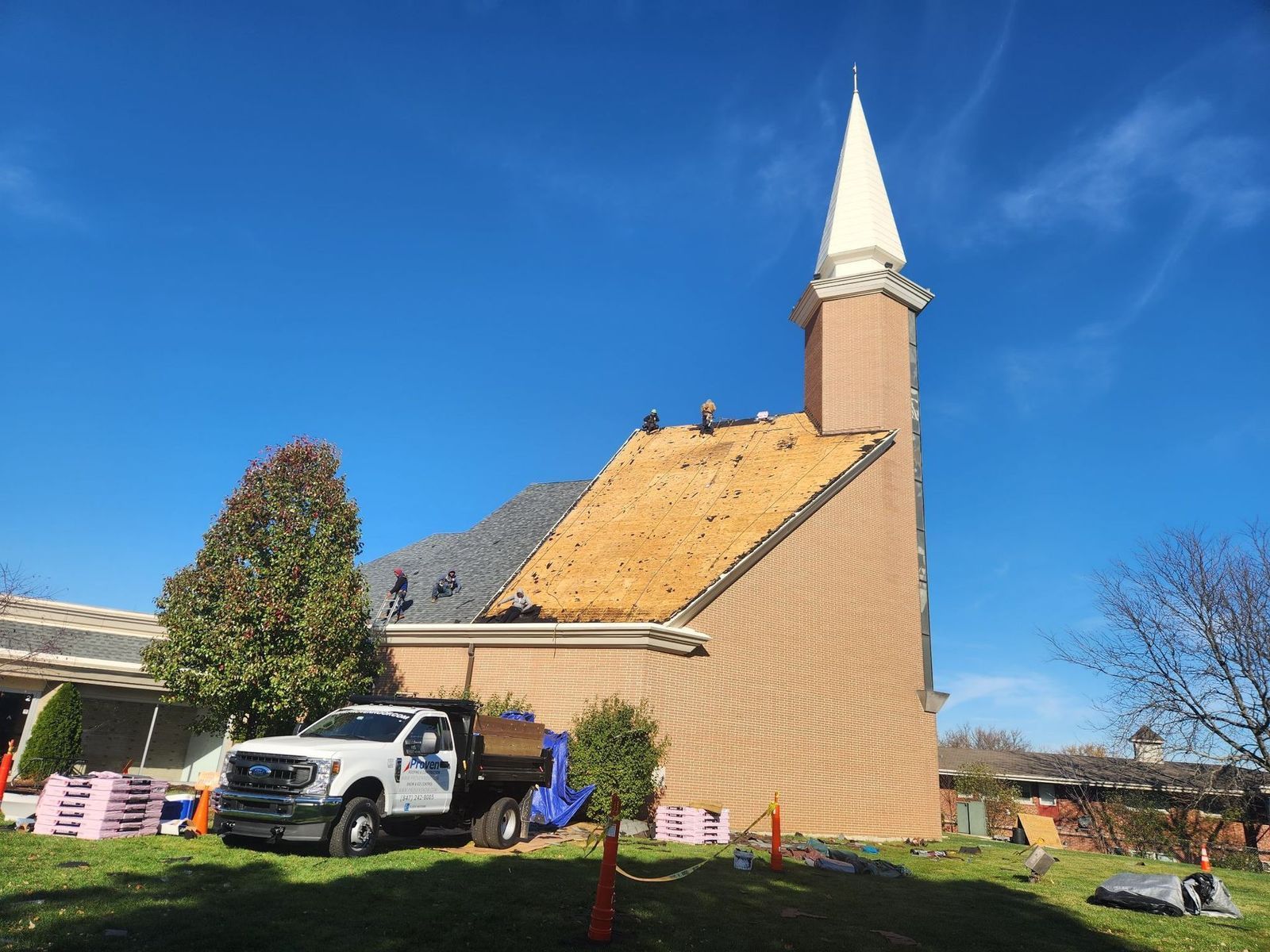 Truck parked near a church with roof repairs; blue sky, brick building, white steeple, brown roof.