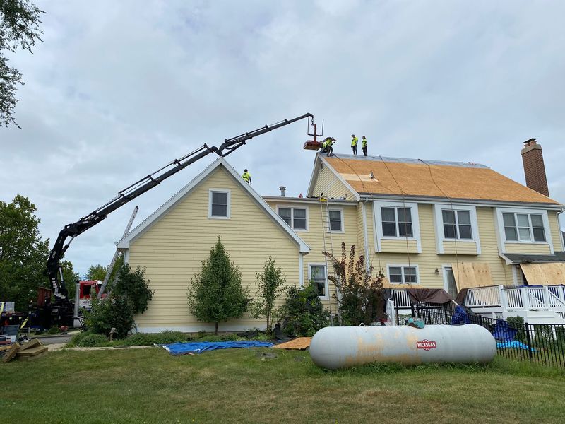 Crane lifts materials to workers on roof of yellow house; propane tank in yard.
