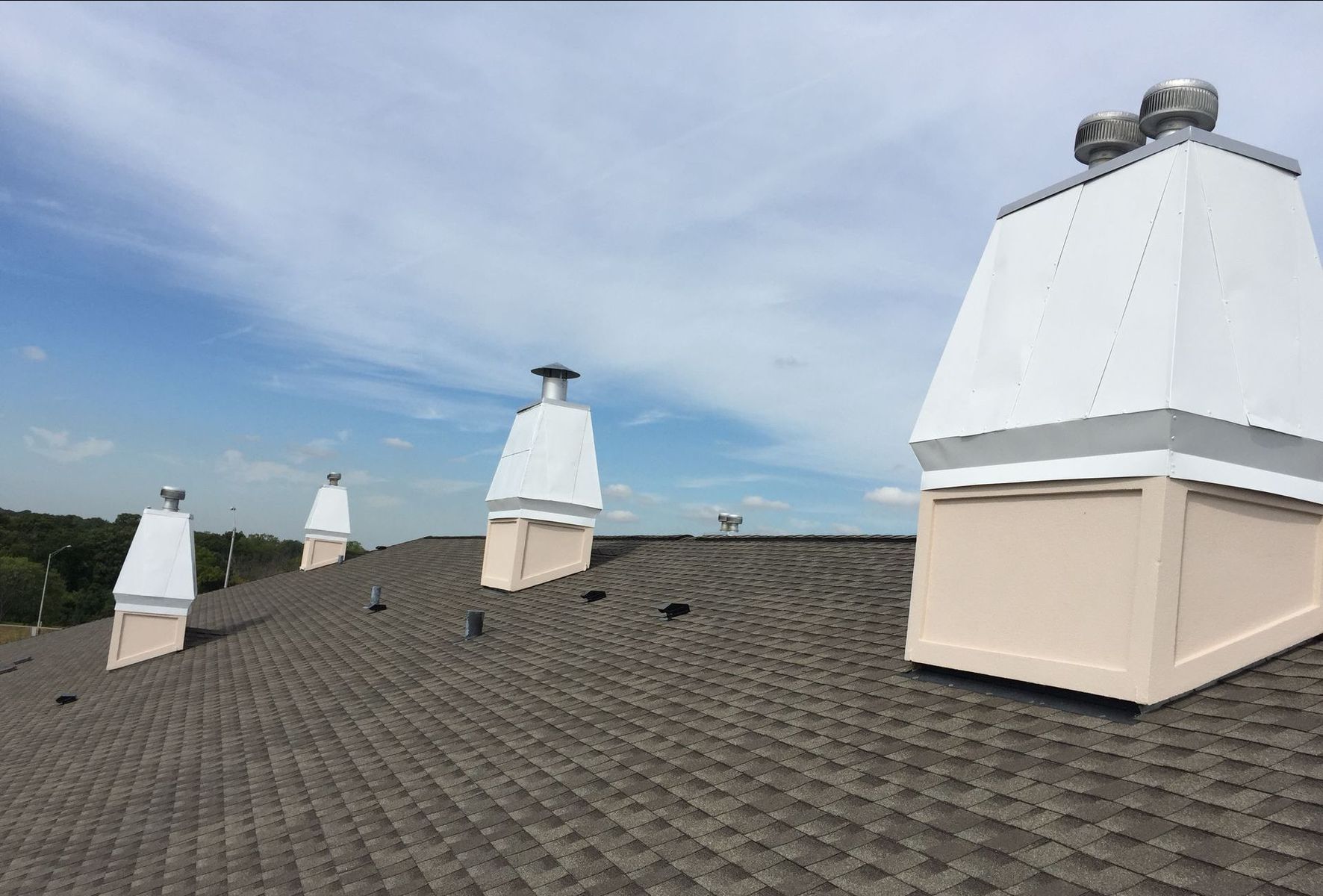 Asphalt shingle roof with four chimneys. White metal chimney caps and pale pink bases against a blue sky.