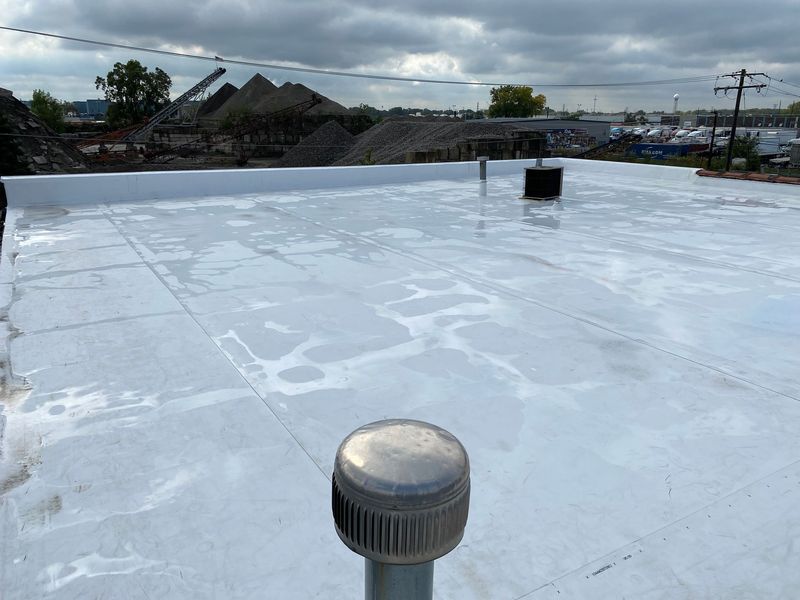 A flat white roof with some water puddles, a vent, and a cloudy sky in the background.