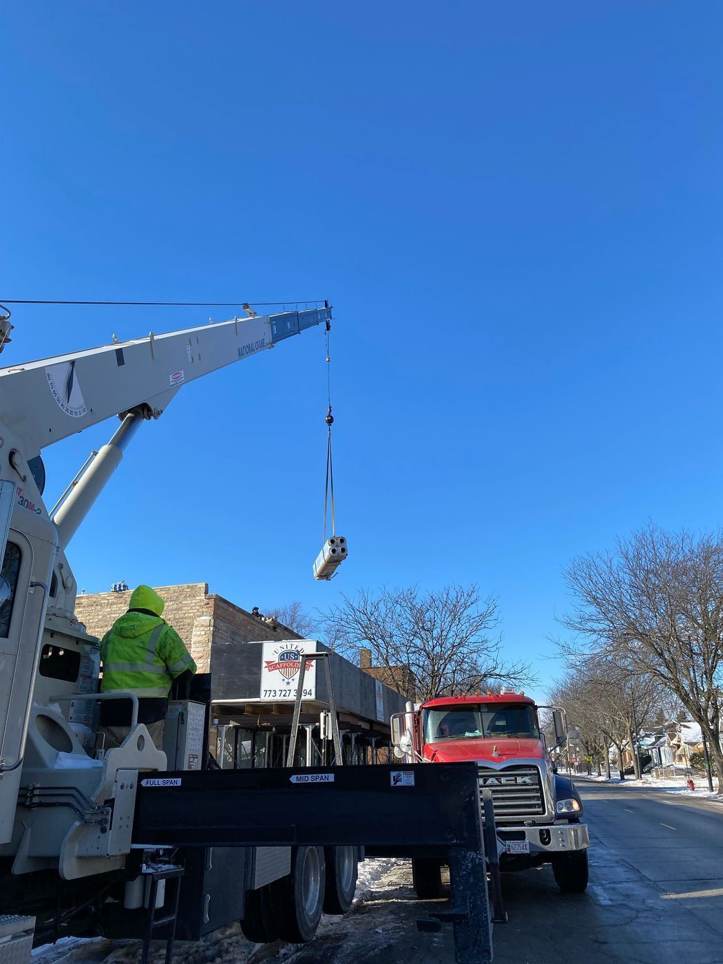 A crane lifting an object in front of a building on a clear, blue sky day. A worker in a green vest is in the crane's cab.