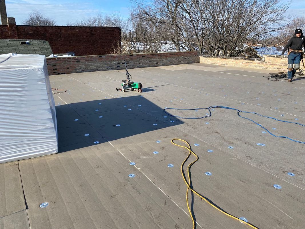 A green robot on a rooftop with a person, blue and yellow ropes, and stacks of building materials on a sunny day.